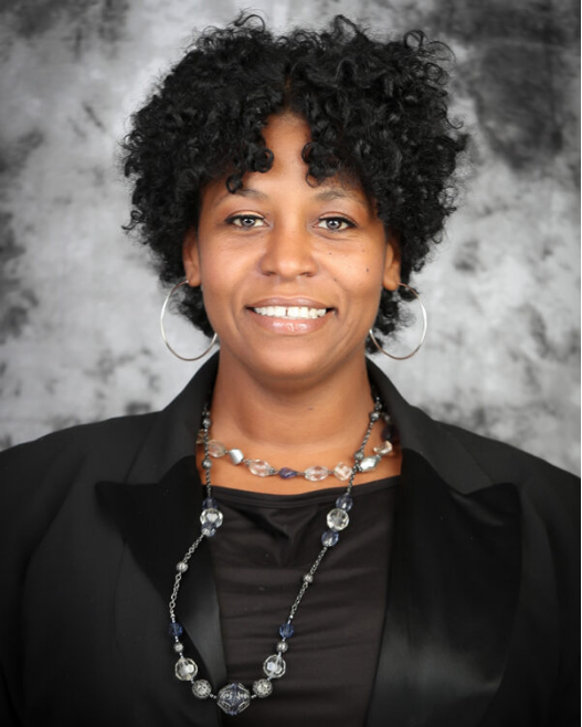 Taryn smiles. She is wearing a black shirt and jacket with necklaces and earrings and has short curly hair.