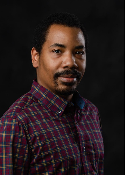 Malcolm looks straight ahead. He is wearing a red, blue, and yellow plaid button-up dress shirt.